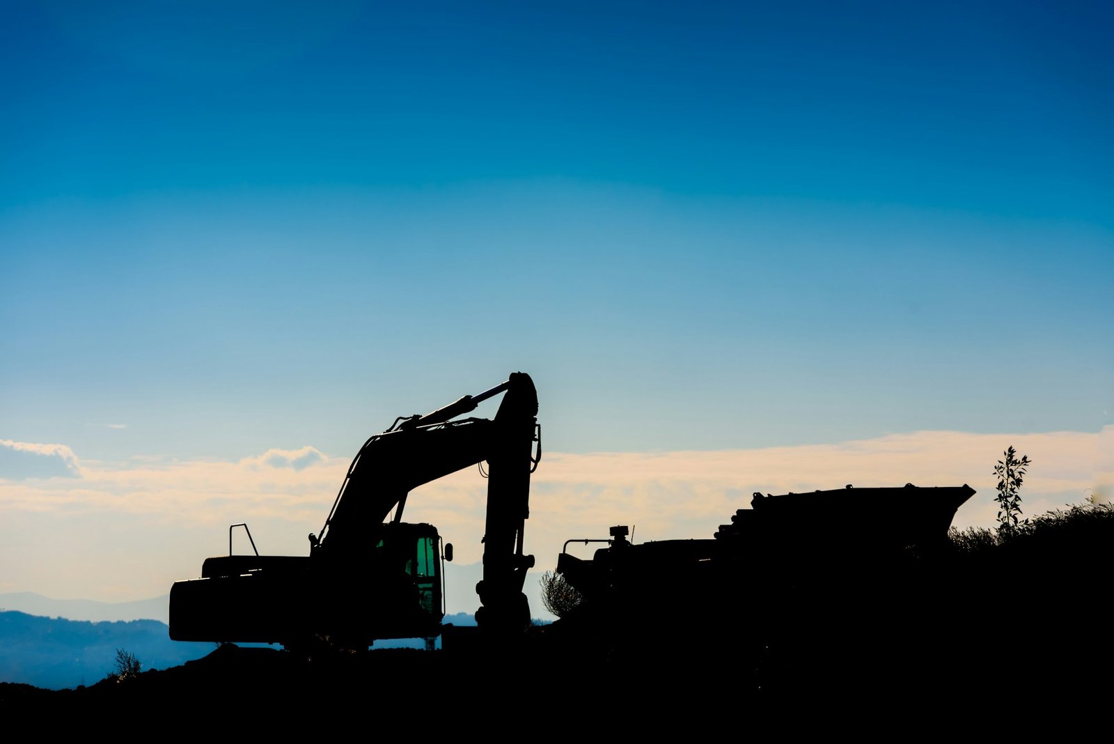 Backlit silhouette of an excavator on top of a hill with unfocused sky background.